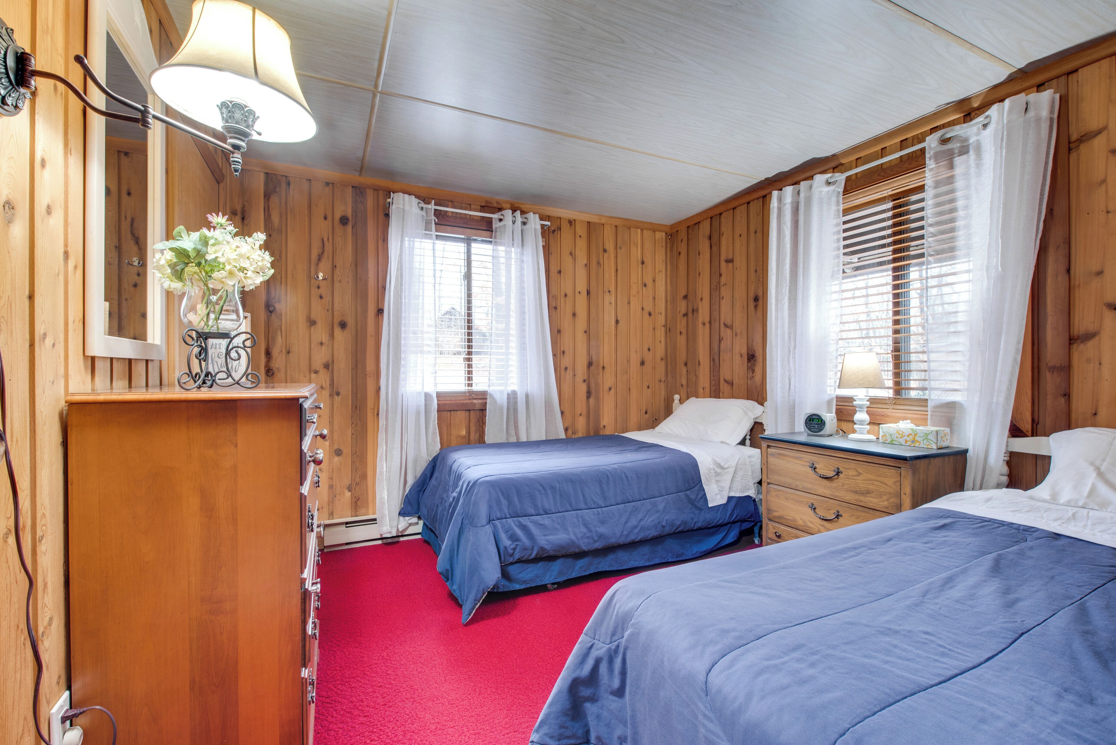 A room with wooden walls, two beds with blue bedding, a red carpet, a wooden dresser, a mirror, and a lamp.