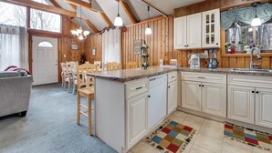 A kitchen with wooden cabinets, a dining area with chairs, and a large window. - Fire Pit + Community Perks: Gouldsboro Cabin! (Gouldsboro)