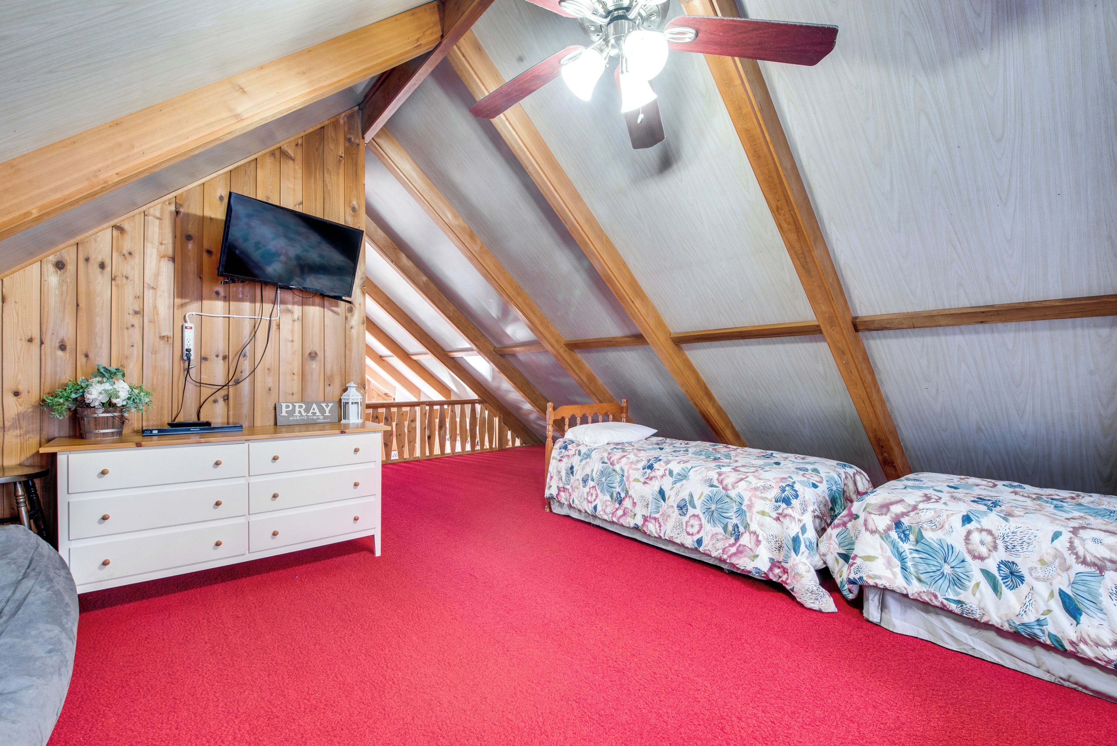 A bedroom with a sloped ceiling, a television, a dresser, two beds, and a red carpet.