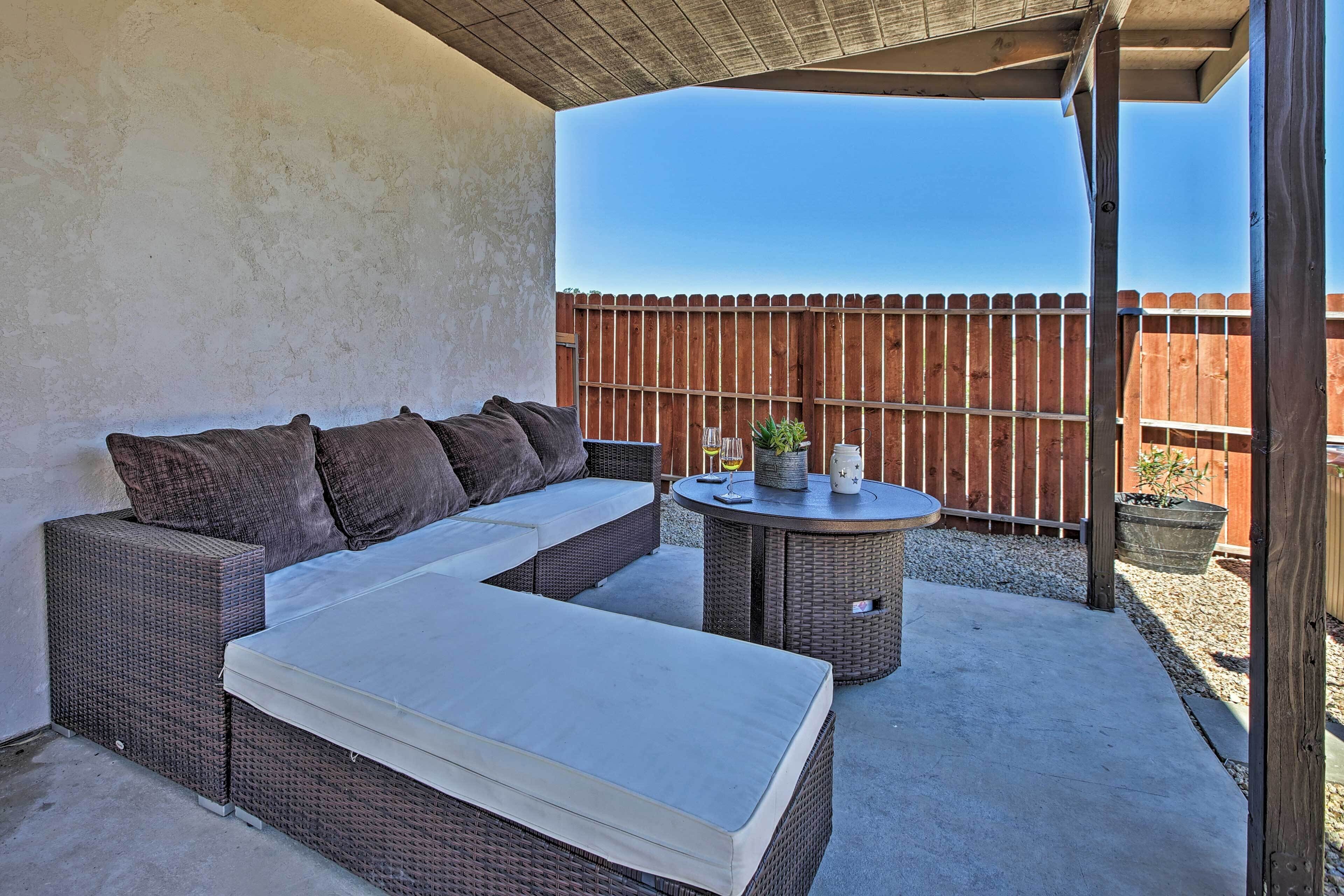 A covered patio area with a wicker sofa and a round table, a wooden fence in the background, and a clear sky.