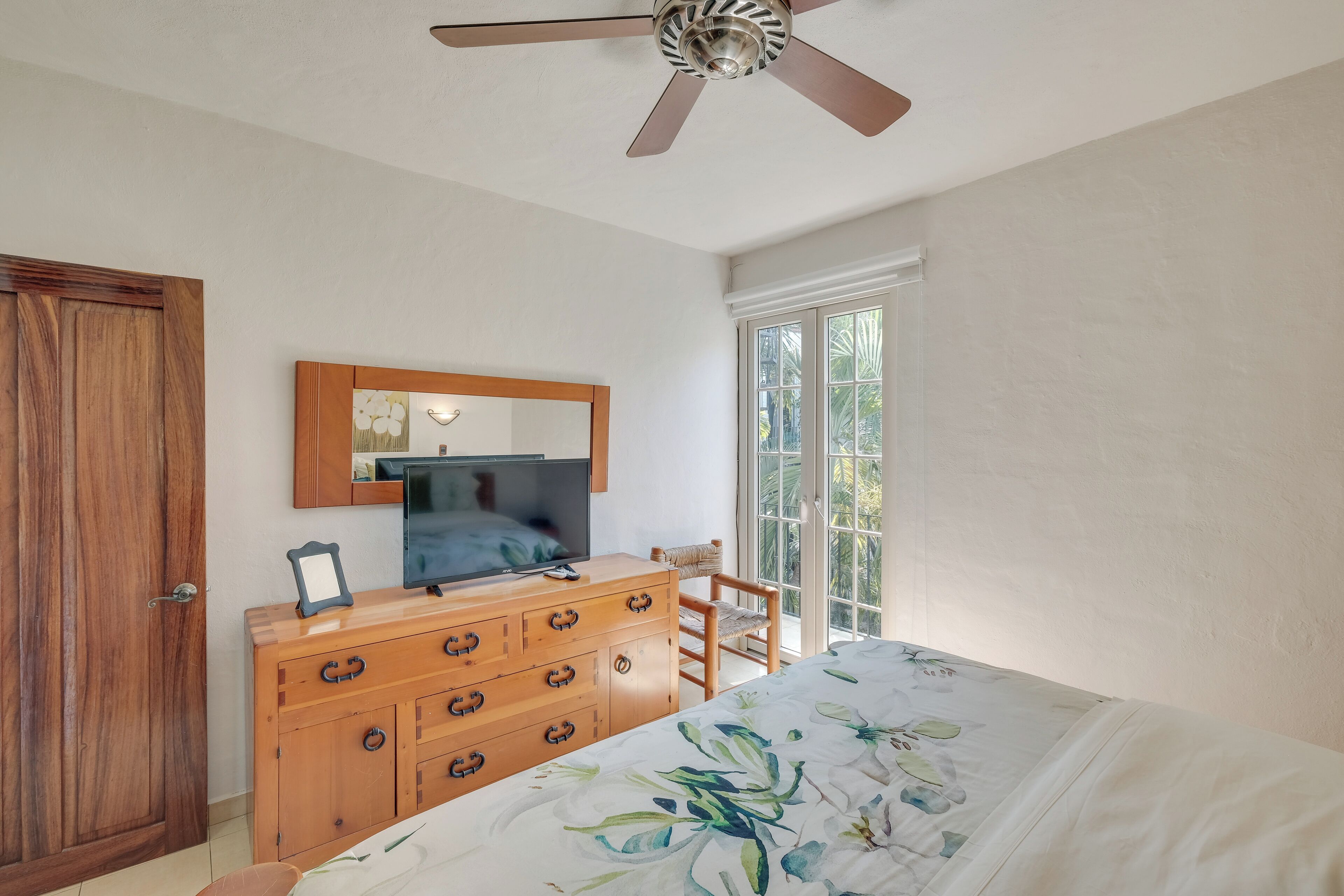 A bedroom with a wooden dresser, a television, a ceiling fan, a window with a view of greenery, and a bed with a floral bedspread.