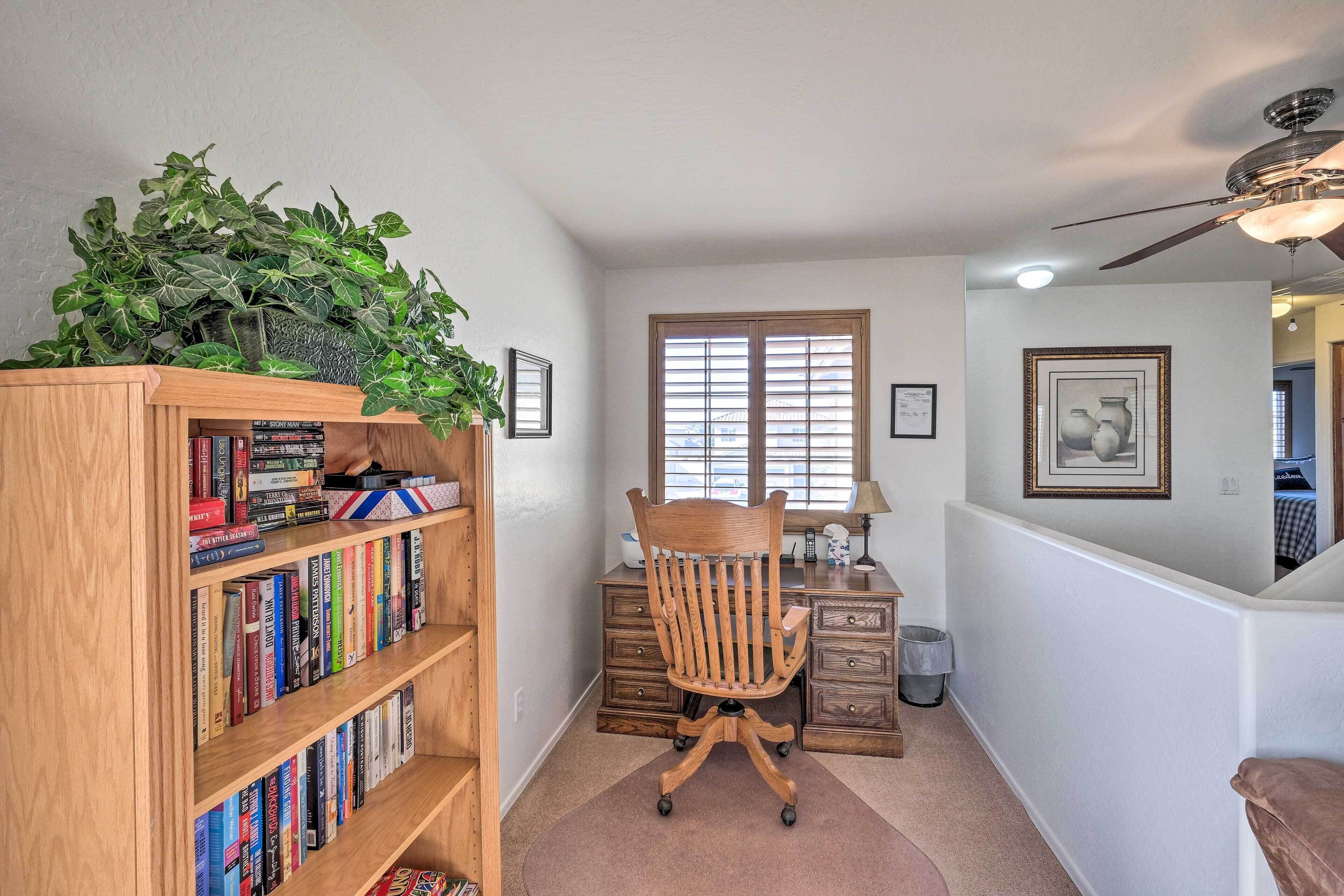 A home office with a bookshelf, a desk, a chair, a window with blinds, and a ceiling fan.