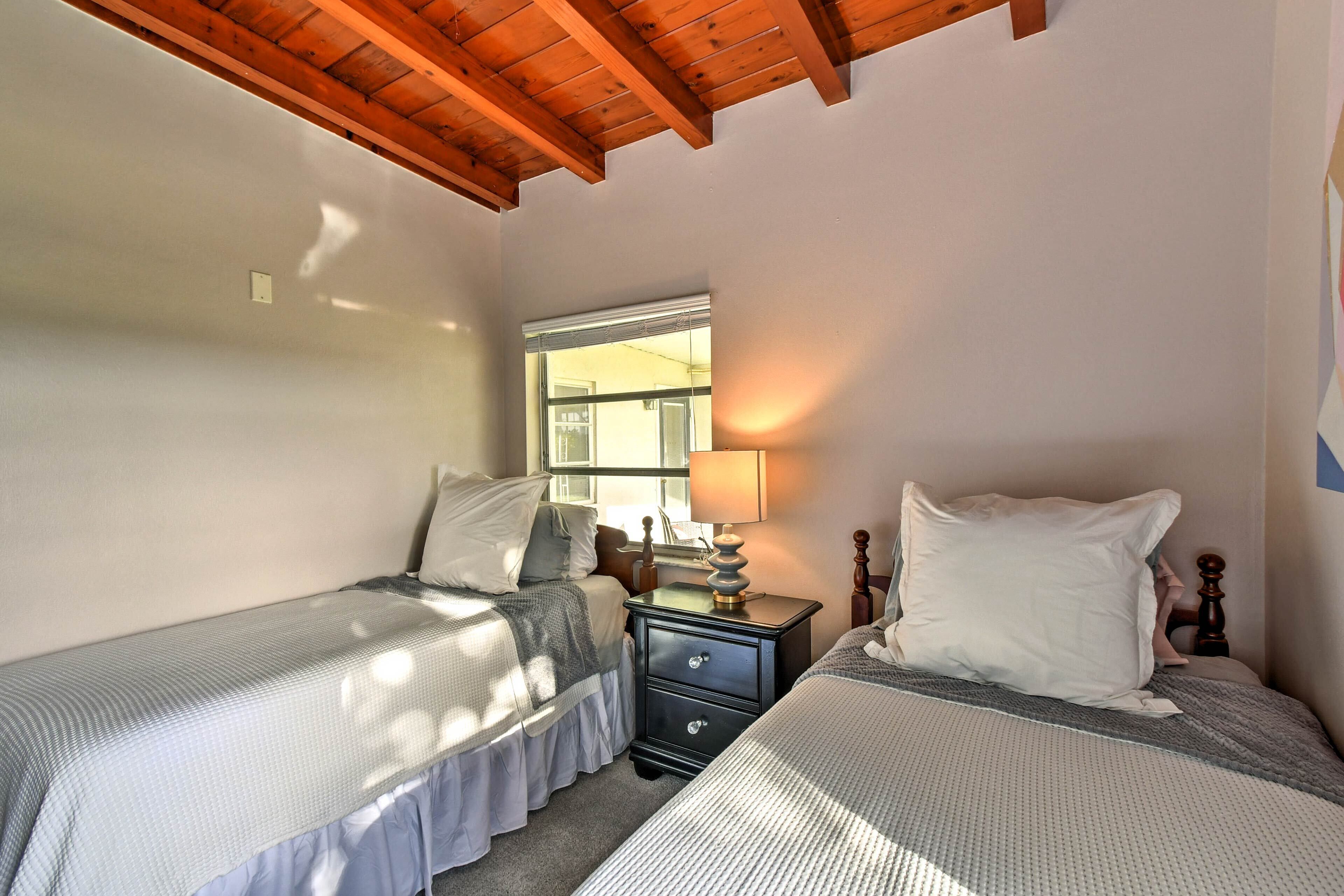 Un dormitorio con dos camas individuales, cabecero de madera, una mesita de noche con lámpara y ventana que proporciona luz natural.