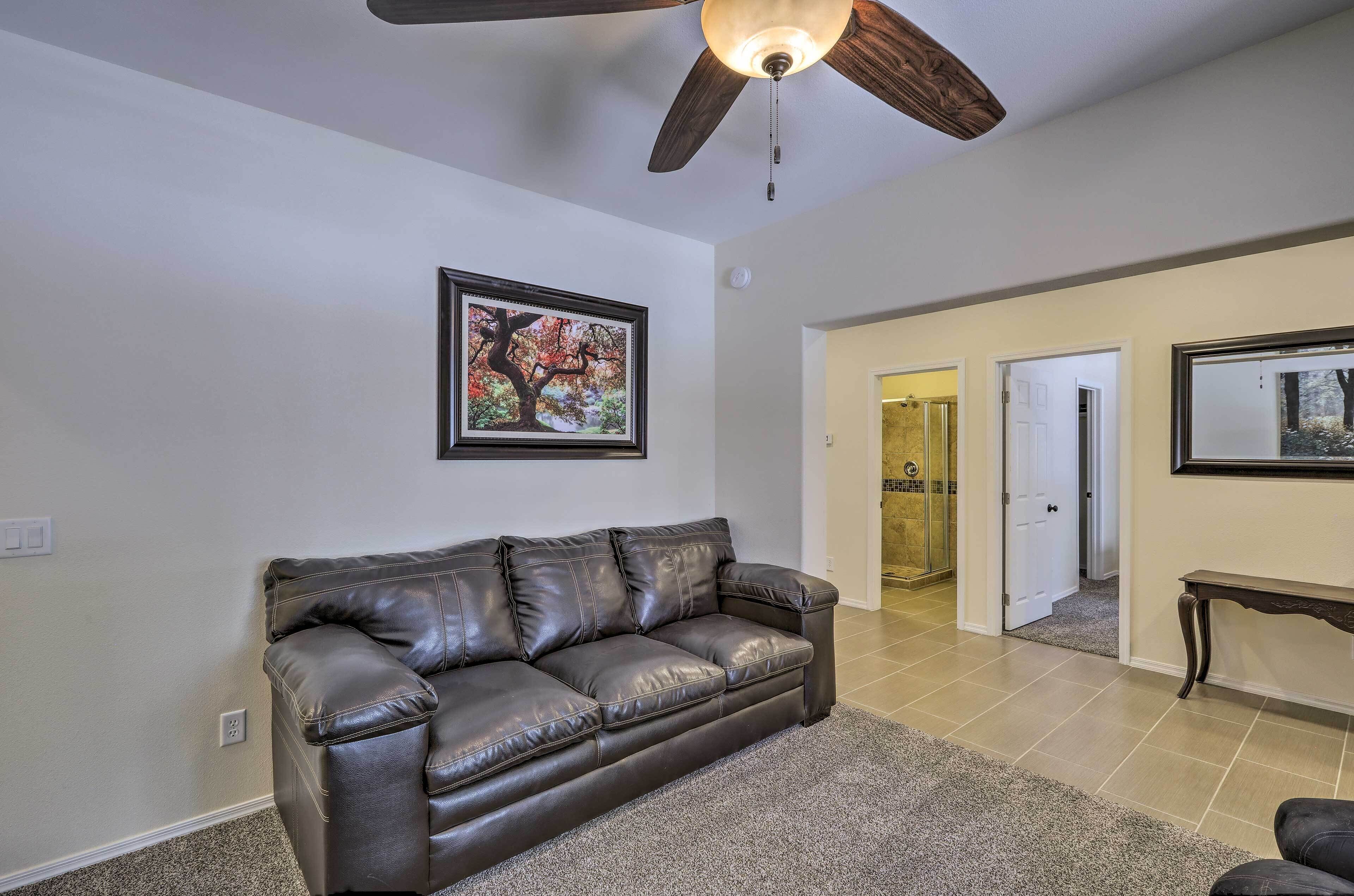 A living room with a dark leather couch, a ceiling fan, a framed picture on the wall, and a hallway leading to a bathroom.