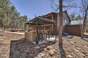 A rustic cabin with a covered outdoor seating area, surrounded by trees and a rocky landscape. - 3 Mi to Rainbow Lake: Home w/ Hot Tub & Deck (Lakeside)