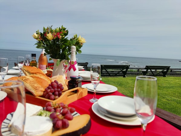 A table set for a meal with a view of the ocean, including plates, glasses, bread, grapes, and a bottle of champagne.