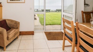 A living room with a sofa, a dining table with chairs, a glass door leading to a balcony with a view of the sea, and a framed picture on the wall. - On The Watersedge (Chathill)