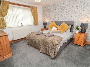 A bedroom with a grey upholstered bed, a fluffy rug, a wooden nightstand, a bedside table, a radiator, a window with curtains, and a floral wallpaper.
