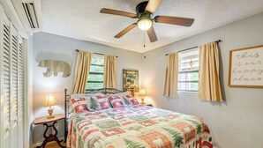 A bedroom with a bed, a ceiling fan, a window with curtains, a bedside table with a lamp, and a wall decoration.