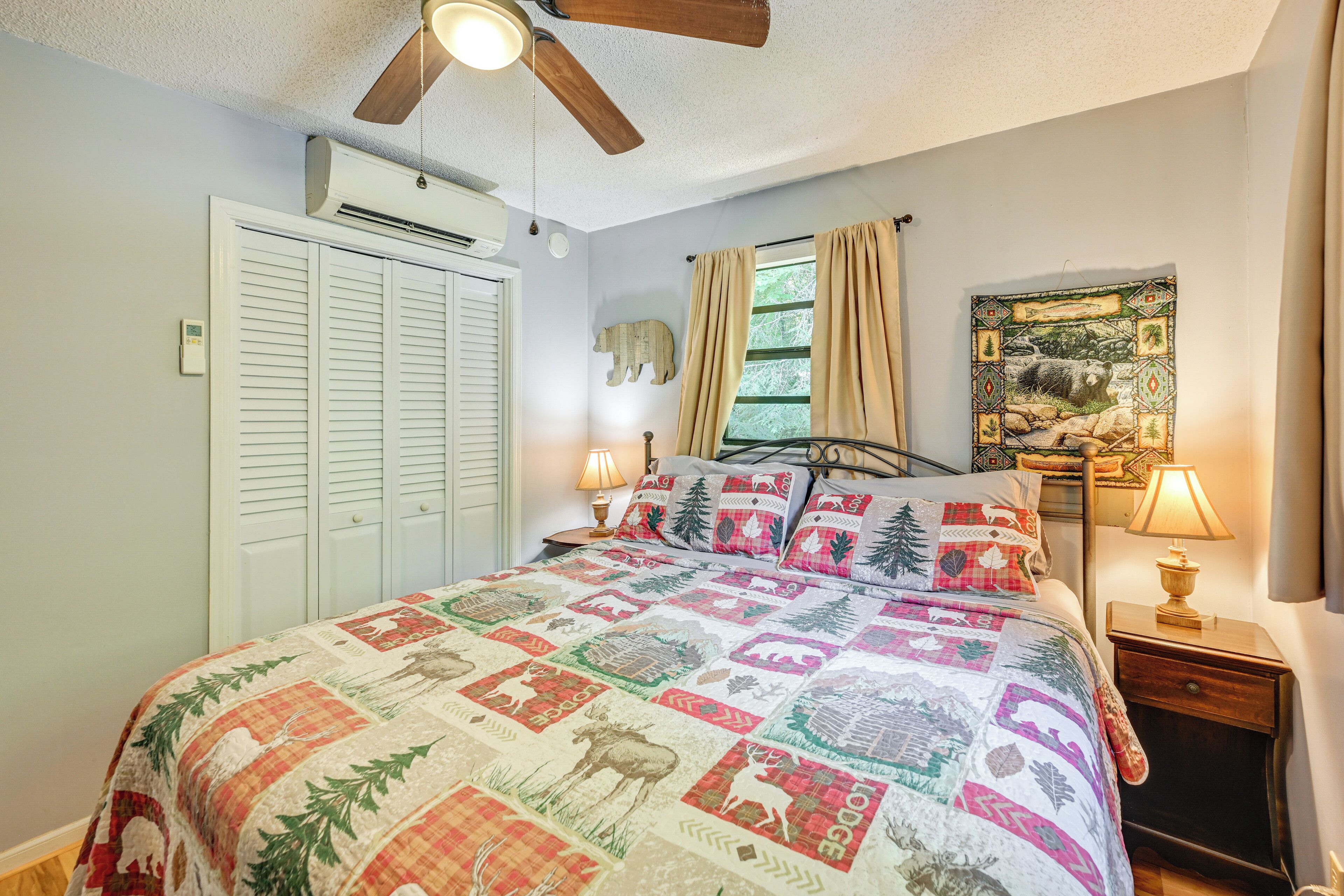 A bedroom with a quilted bedspread, a ceiling fan, a window with curtains, a bedside table with a lamp, and a wall-mounted air conditioner.