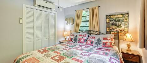 A bedroom with a quilted bedspread, a ceiling fan, a window with curtains, a bedside table with a lamp, and a wall-mounted air conditioner.
