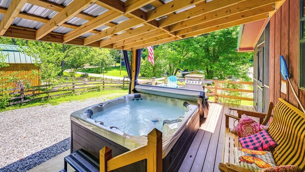 A hot tub under a wooden pergola with a wooden deck, surrounded by a wooden fence and greenery.