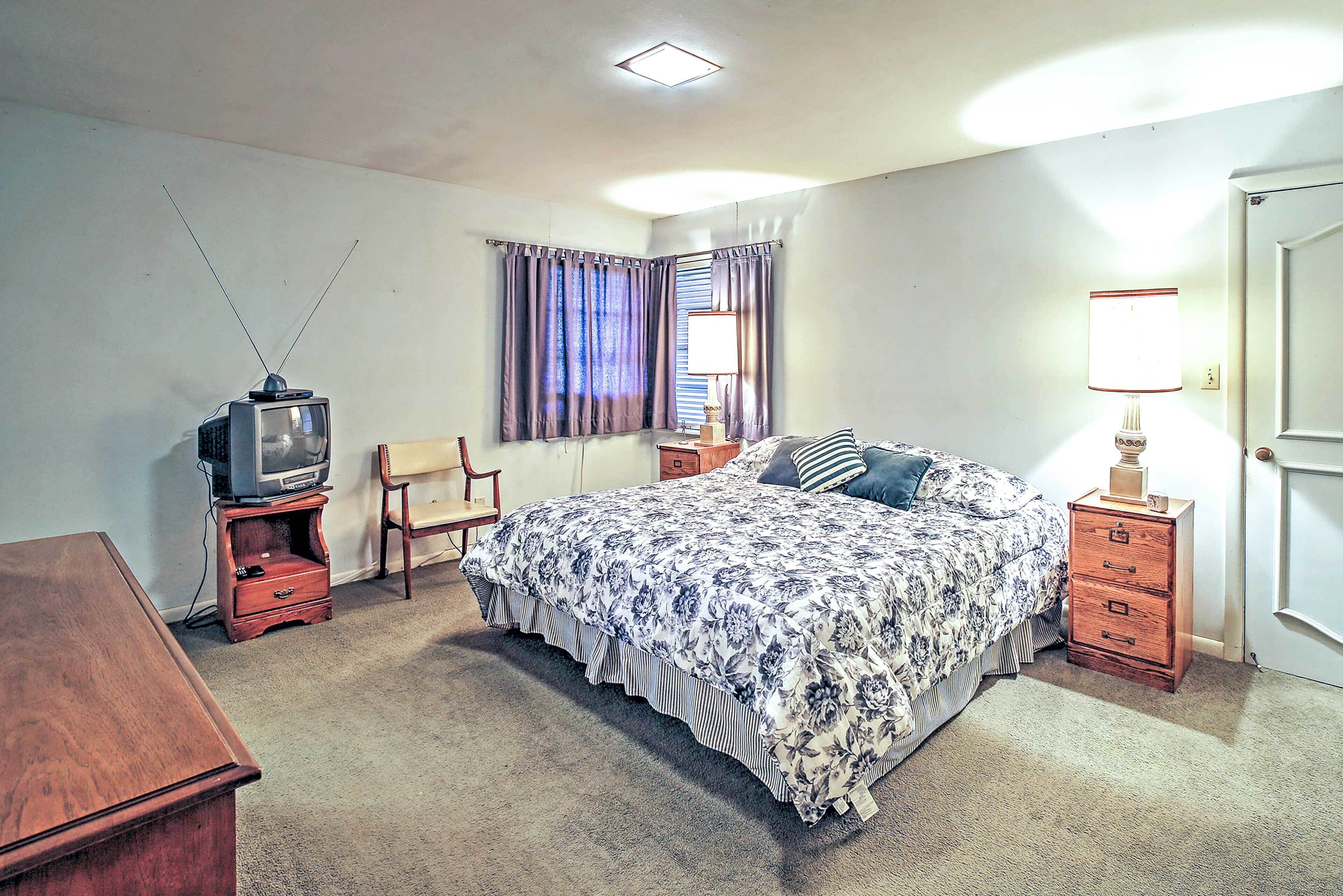 A bedroom with a bed, a television on a stand, a chair, a nightstand, and a lamp.
