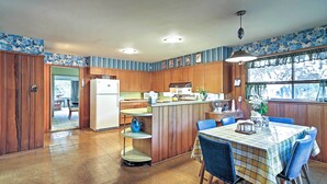 A kitchen with wooden cabinets, a dining table with blue chairs, and a window with a view of trees. - Mid-century Hideaway: Spacious Brownsville Home! (Brownsville)