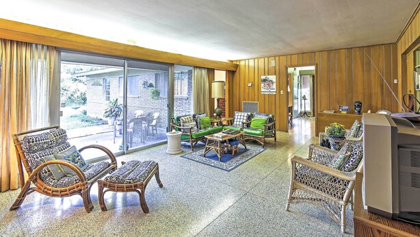A spacious living room with a large sliding glass door leading to a patio, wicker furniture, a television, and a wooden paneled wall. - Mid-century Hideaway: Spacious Brownsville Home! (Brownsville)