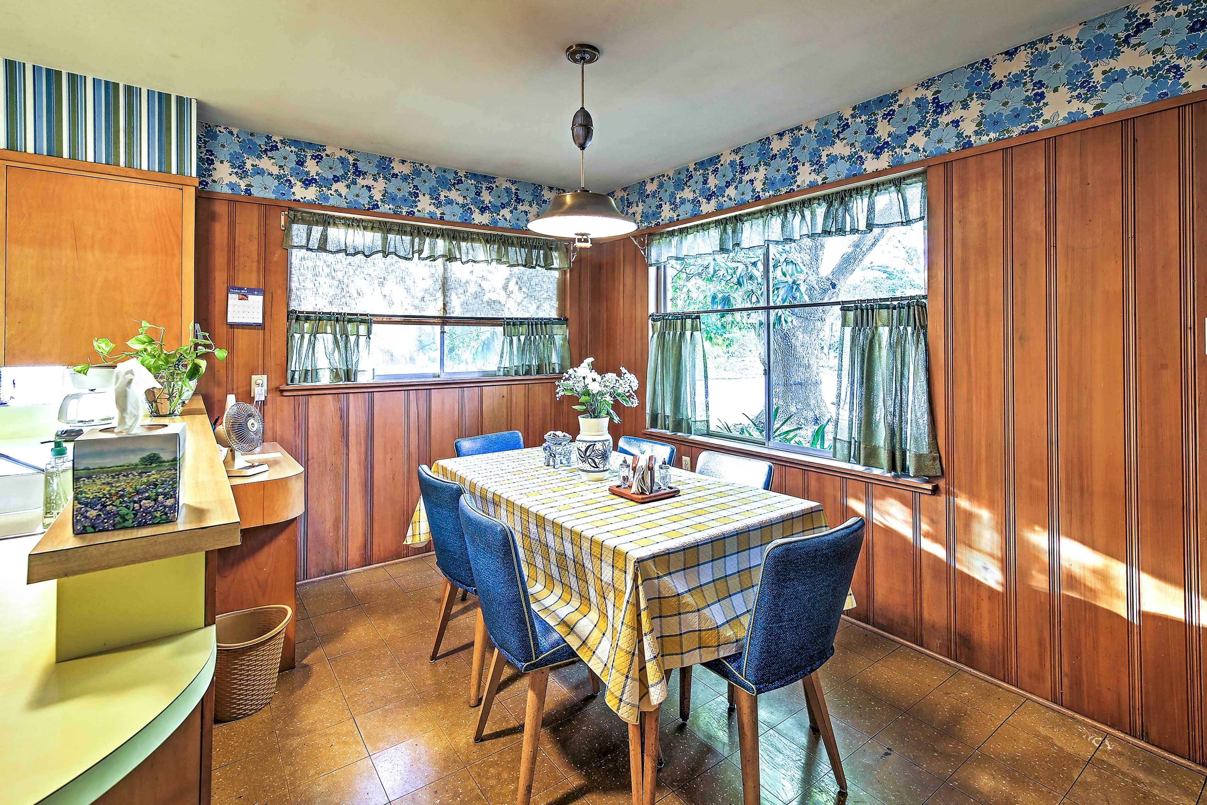 A dining area with a wooden table, blue chairs, and a checkered tablecloth. There is a window with curtains and a view of greenery outside.