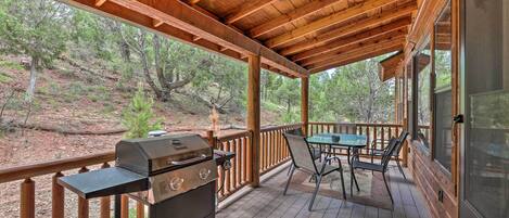 A covered outdoor deck with a grill and table, surrounded by wooden railings and overlooking a wooded area.