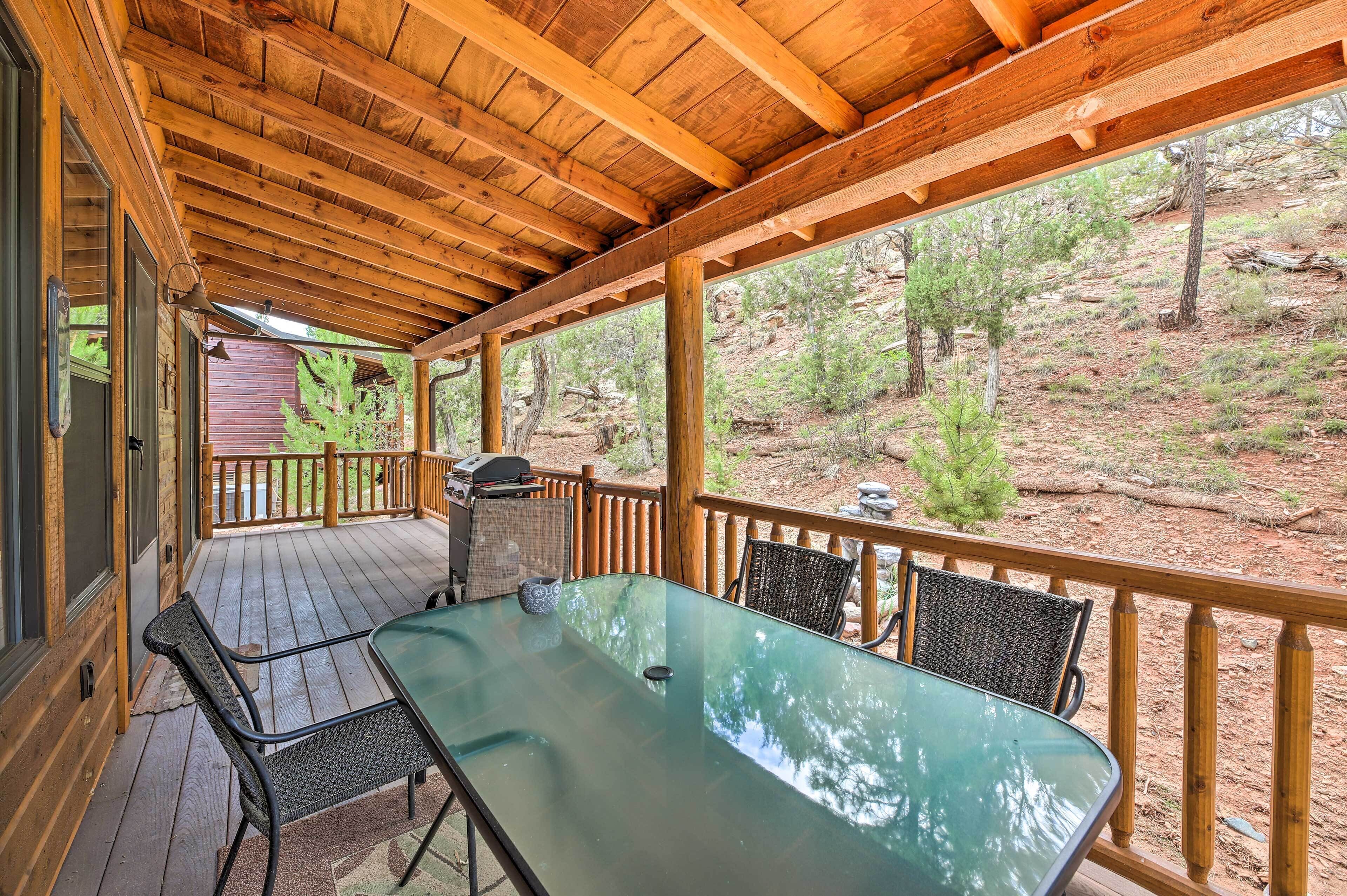 Un patio cubierto al aire libre con mesa y sillas de vidrio, barandales de madera y vistas a un área boscosa.