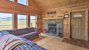 L’intérieur chaleureux d’un chalet en bois, avec une cheminée, un canapé à carreaux et de grandes fenêtres donnant sur le paysage extérieur.