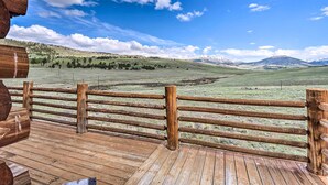 Une terrasse en bois, entourée d’une clôture également en bois, donnant sur un champ verdoyant avec des montagnes qui se dessinent à l’horizon.