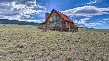 A wooden cabin with a red roof, multiple windows, and a porch, situated in a grassy field with a mountain in the background.