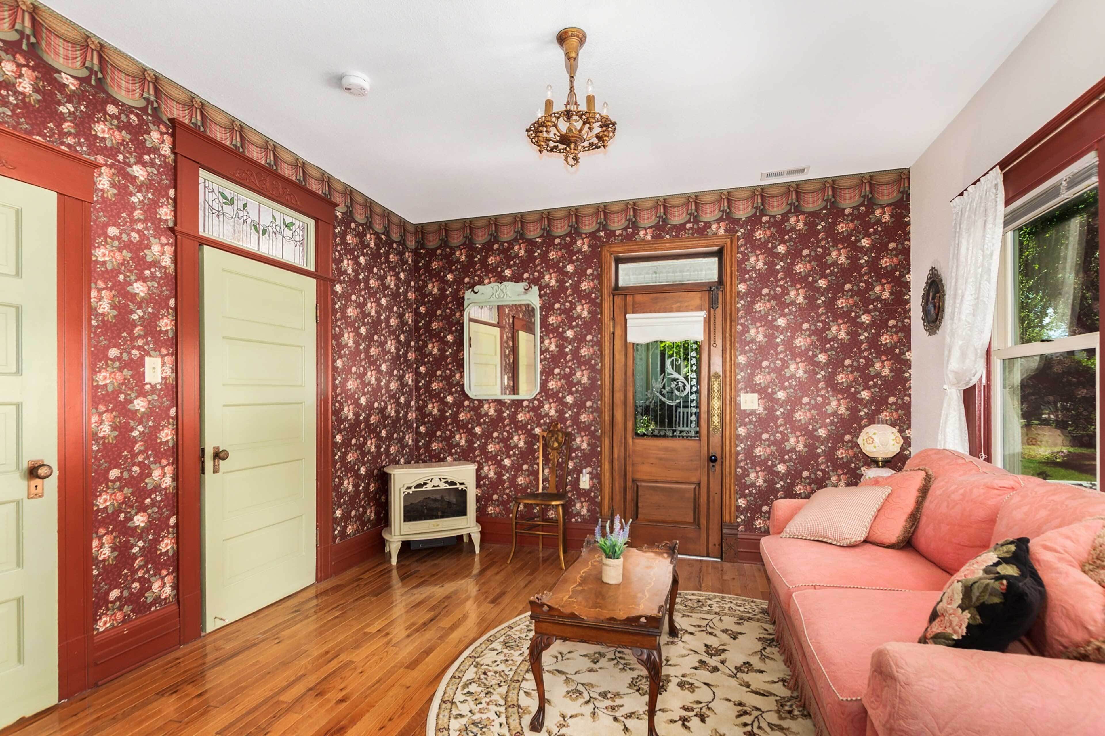A room with floral wallpaper, a wooden floor, a pink sofa, a small table, a mirror, and a chandelier.