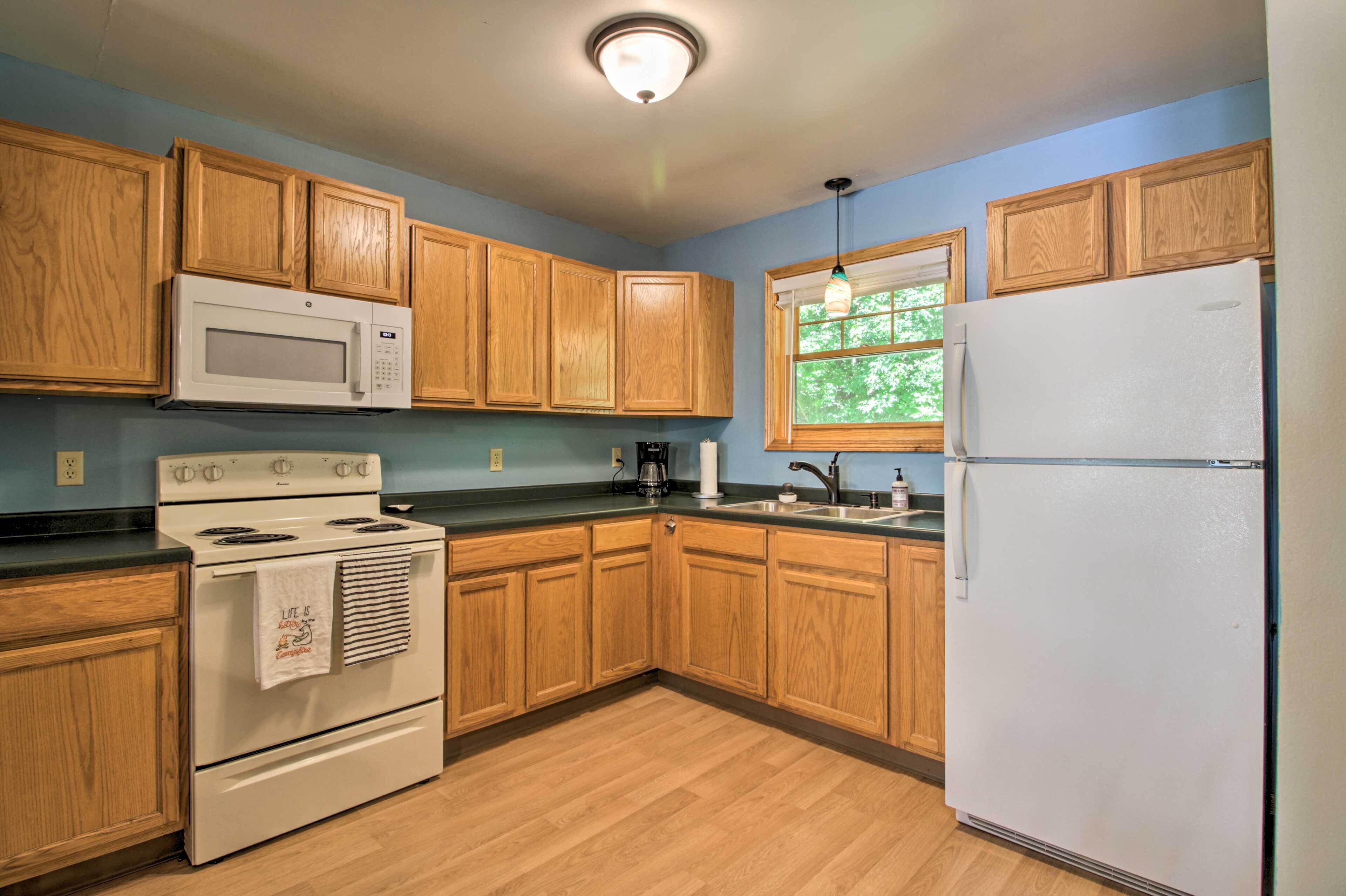 A kitchen with wooden cabinets, a white microwave, stove, and refrigerator, a window with a view of trees, and a light fixture on the ceiling.