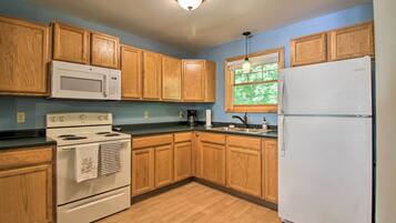 A kitchen with wooden cabinets, a white microwave, stove, and refrigerator, a window with a view of trees, and a light fixture on the ceiling.