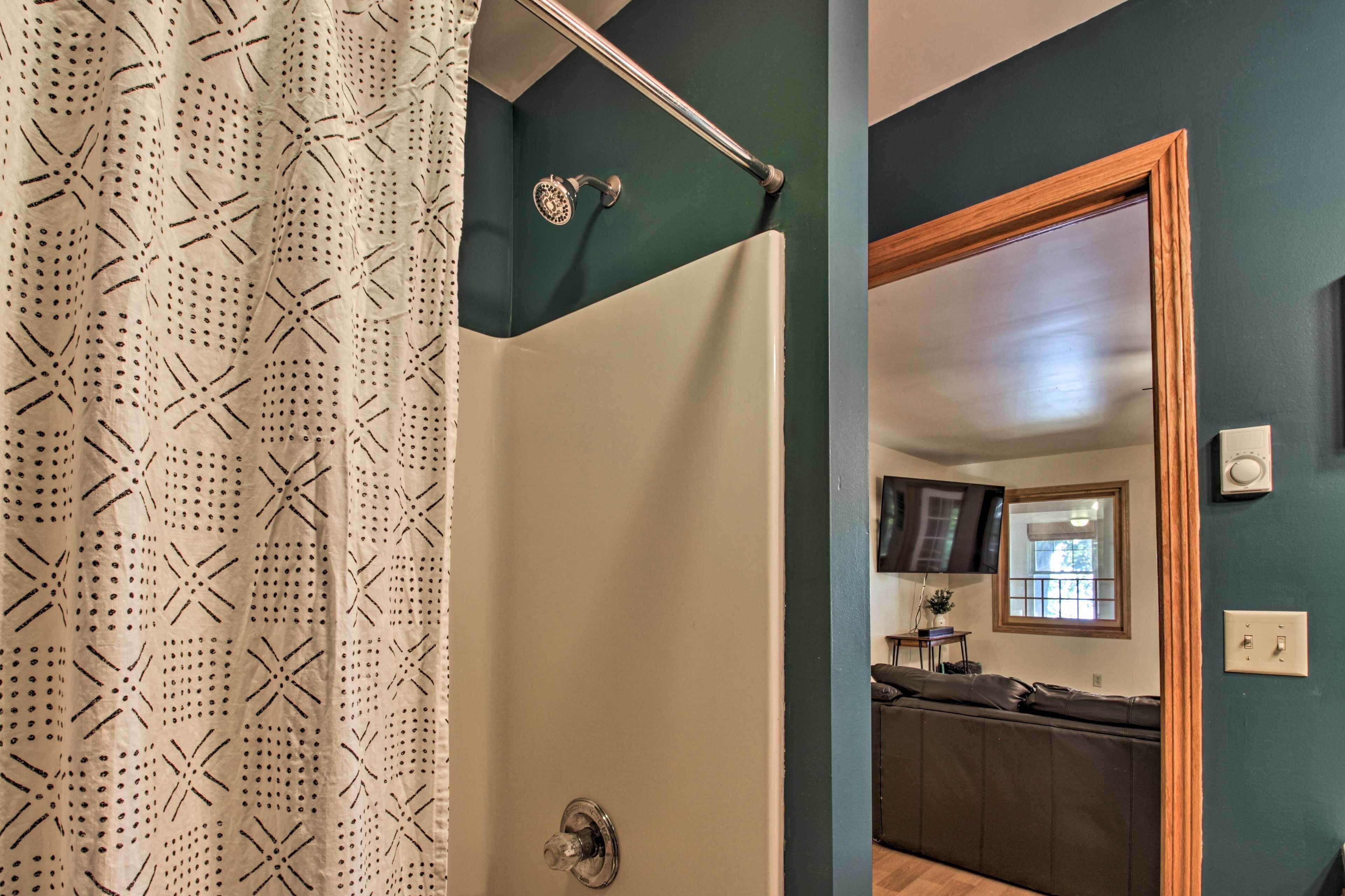 A bathroom with a shower curtain, a bathtub, and a door leading to a living area with a couch and a window.