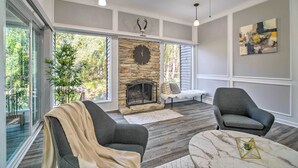 A modern living room with a stone fireplace, two armchairs, a coffee table, and large windows offering a view of greenery. - Coldspring Cottage < 1 Mi to Lake Livingston! (Coldspring)