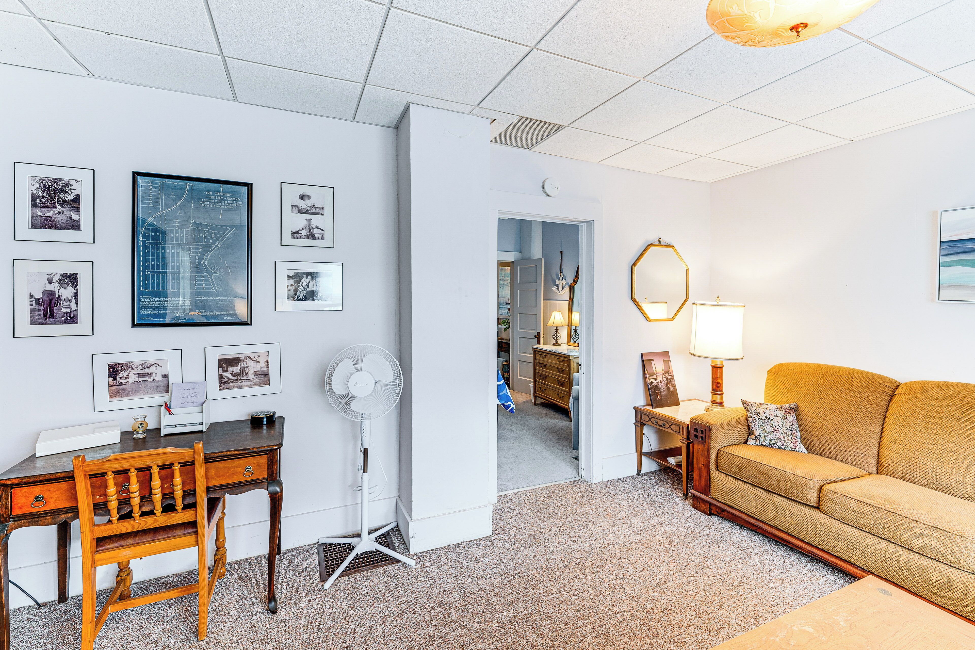 A living room with a wooden desk, a white fan, a sofa, and a wall with framed photographs.