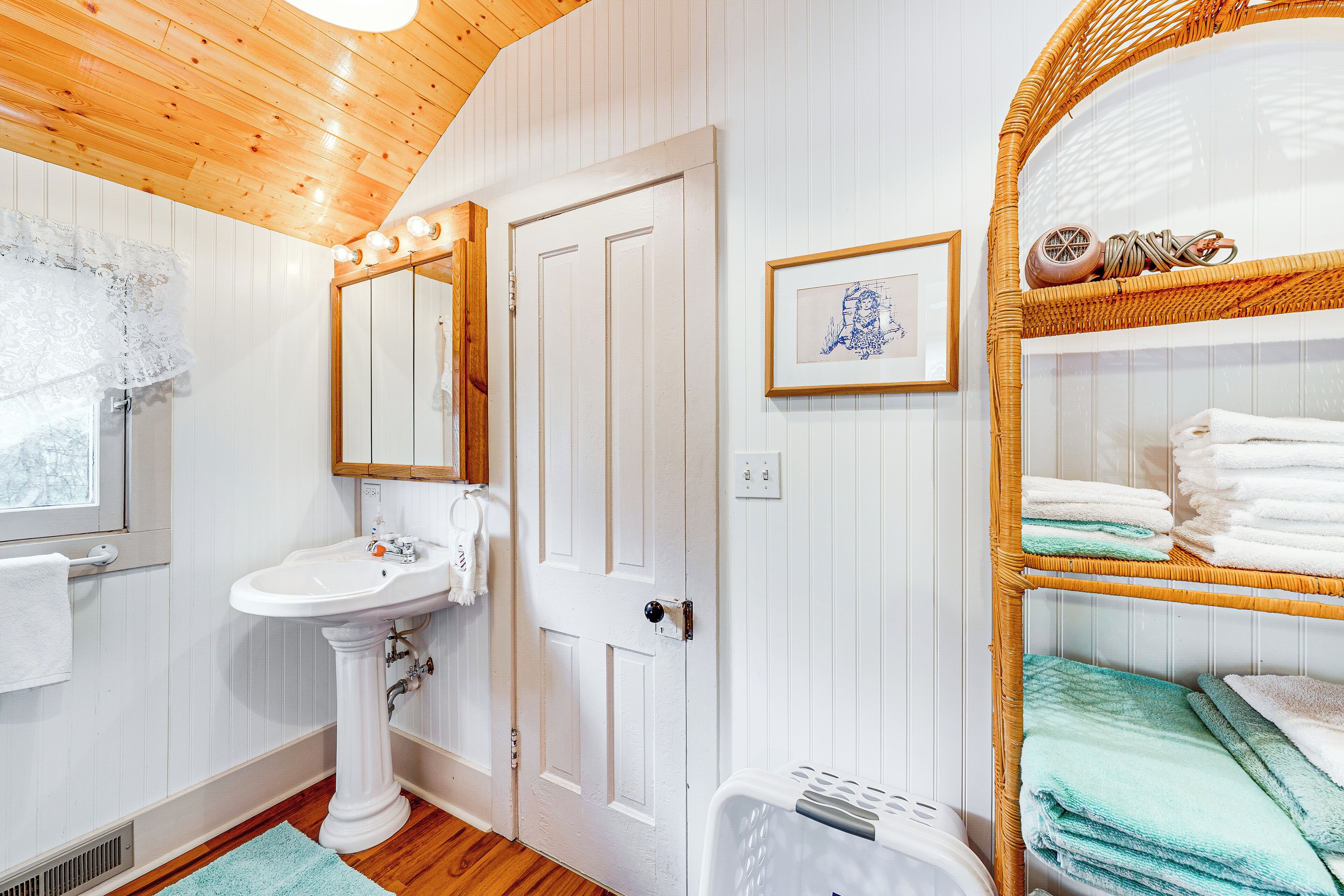 A bathroom with a pedestal sink, a mirror, a door, and a wicker shelf with towels.