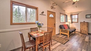 A cozy living room with a wooden dining table, chairs, a sofa, a ceiling fan, and a view of trees through the windows.