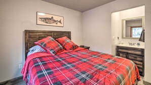 A bedroom with a plaid bedspread, a wooden headboard, a framed fish artwork, a bathroom with a wooden vanity, and a window with a view.