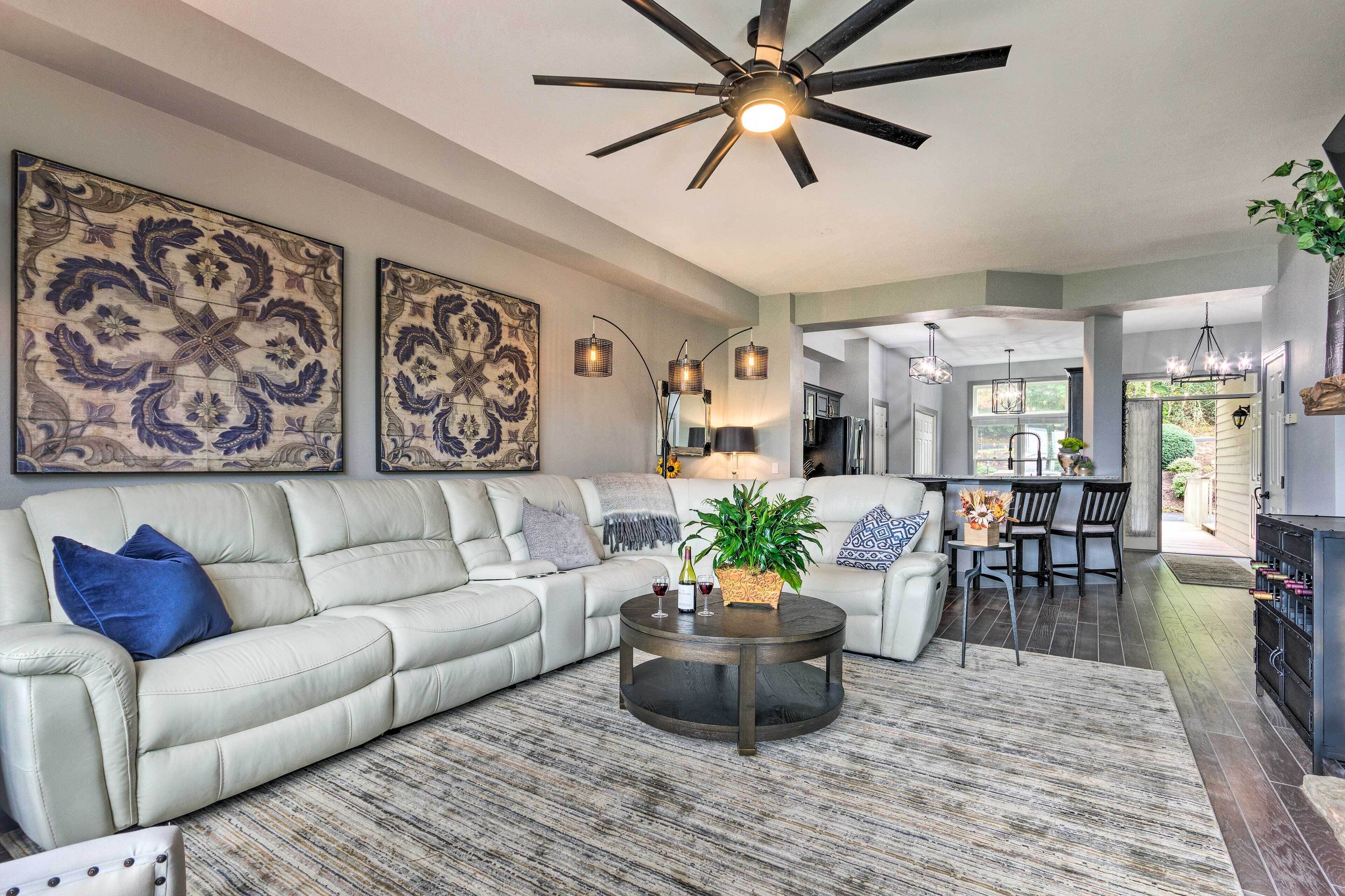 A spacious living room with a sectional sofa, a round coffee table, and a ceiling fan. There are two large wall-mounted artworks and a dining area visible in the background.