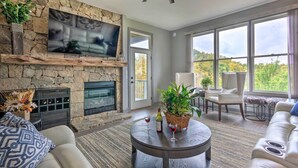 A spacious living room with a stone fireplace, a ceiling fan, and large windows offering a view of the outdoors. - Surrounding Mtn Views! Modern Smokies Retreat (Burnsville)
