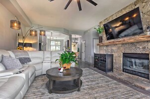 A modern living room with a stone fireplace, a large flat-screen TV mounted above it, a white leather sofa, and a round coffee table with a vase and wine glasses.