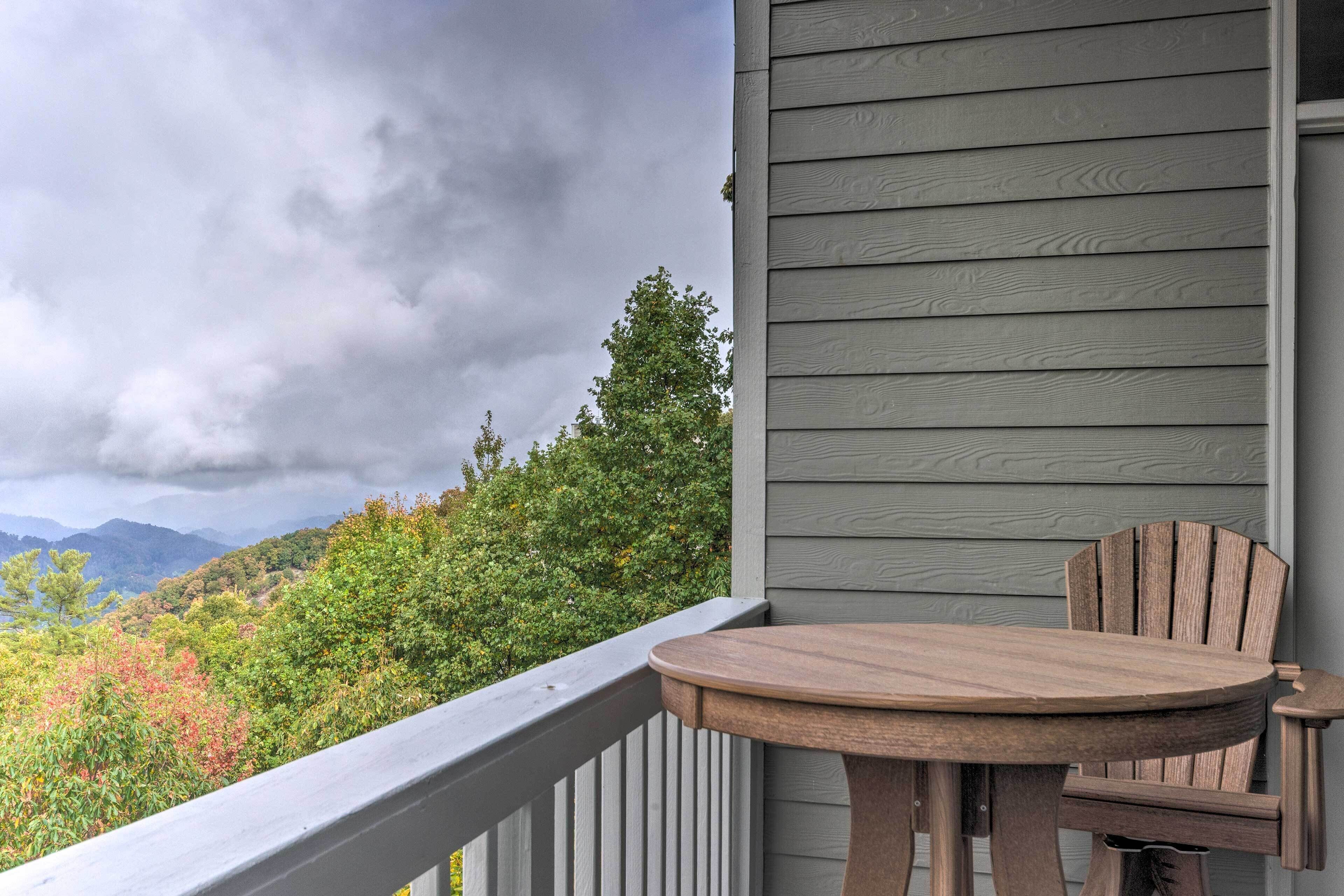 A balcony with a wooden table and chair, overlooking a forested area with mountains in the distance.