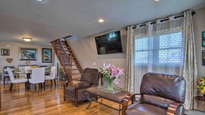 Un amplio salón con una escalera de madera, sillones de cuero, una mesa de comedor con sillas y un gran ventanal con cortinas.