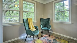 Une pièce avec deux fauteuils verts, un tapis coloré et de grandes fenêtres donnant sur des arbres.