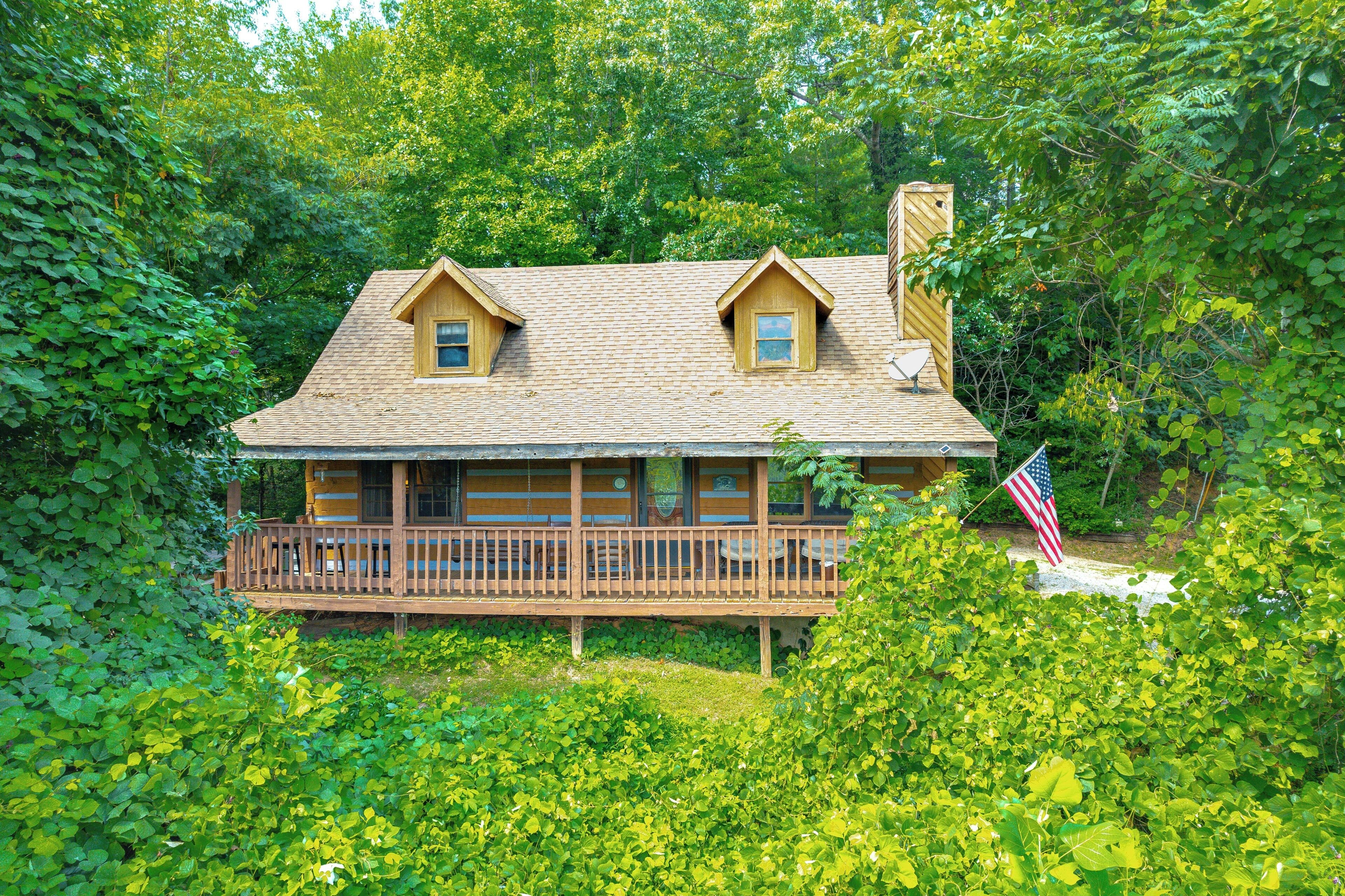 A cabin with a wooden deck and two dormer windows, surrounded by dense green foliage.