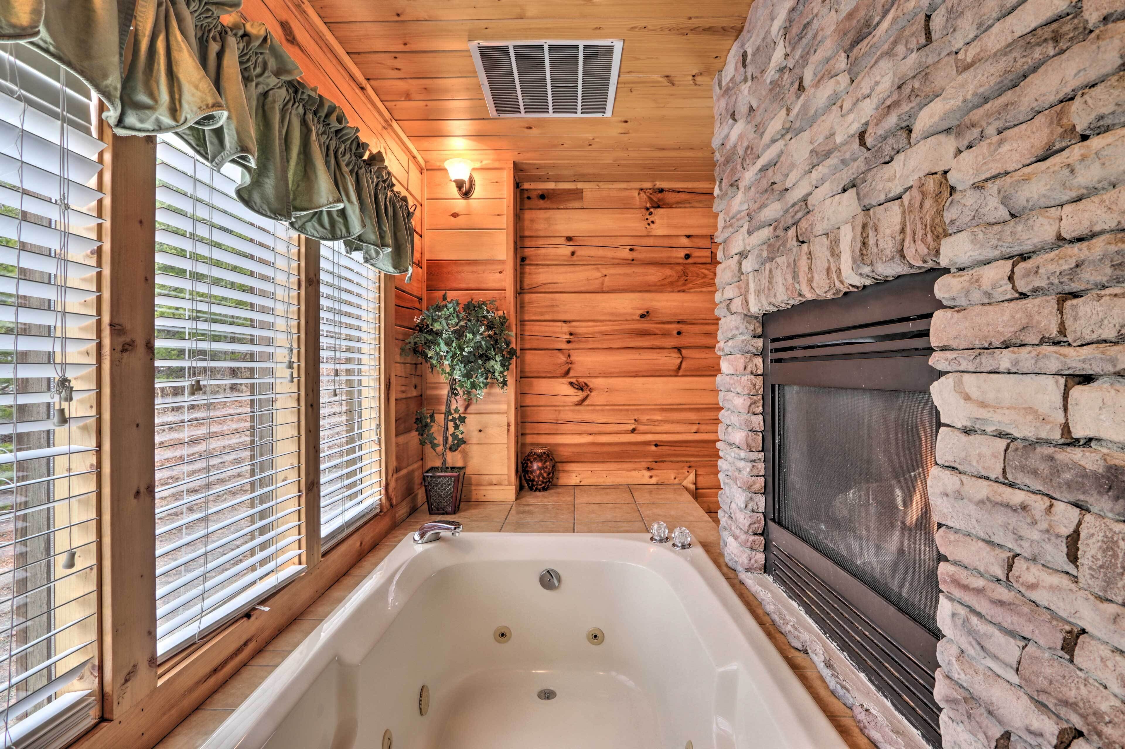 A log cabin bathroom with a large bathtub, stone fireplace, wooden walls, and a window with blinds.