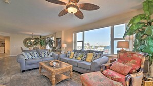 A spacious living room with a large gray sofa, a wooden coffee table, and a red armchair. A ceiling fan and large windows provide natural light.
