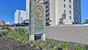 A sign for Twin Towers by Mucci with a pelican logo, surrounded by greenery and a parking area.