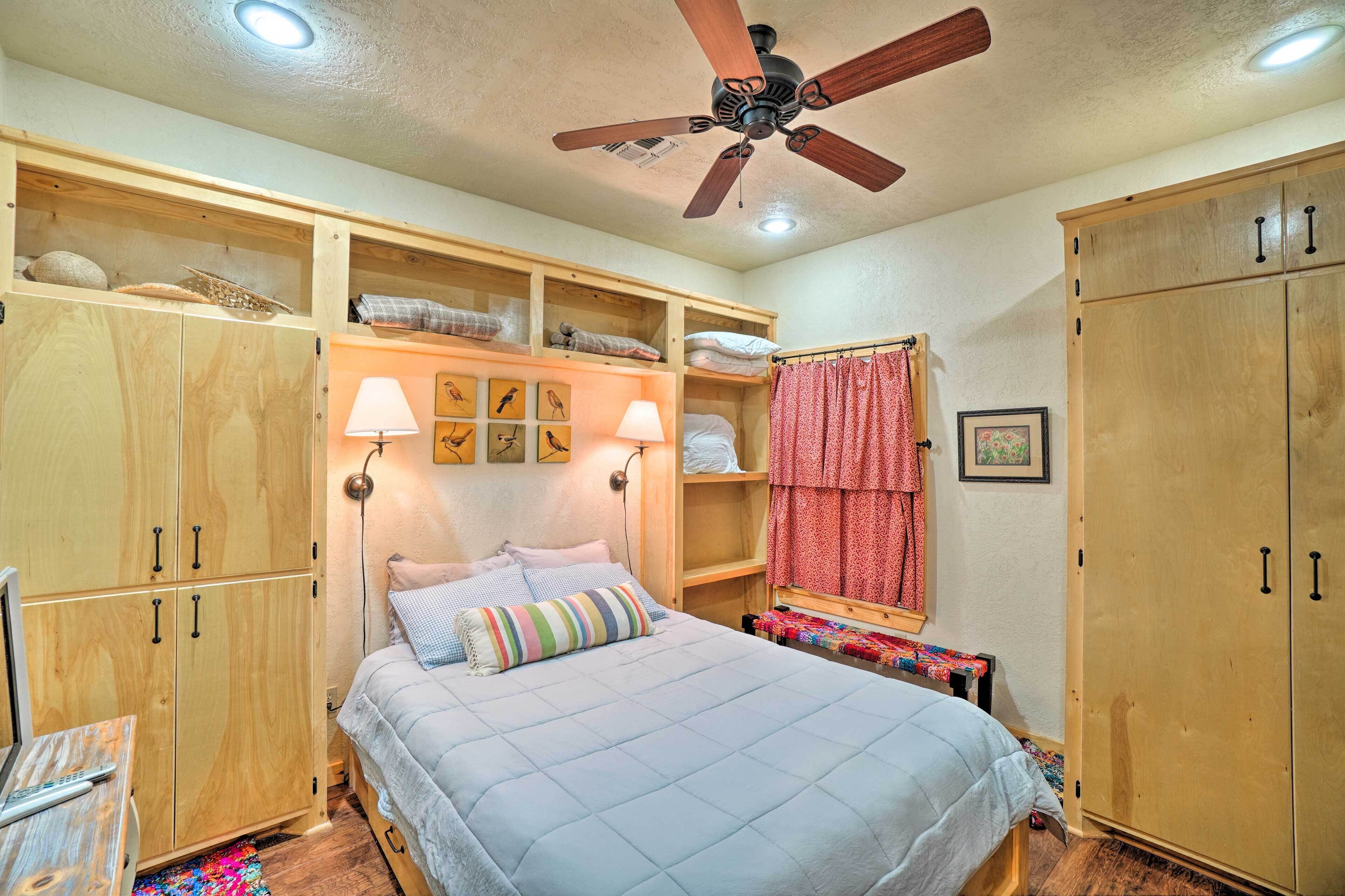 Un dormitorio con cama, armario de madera, estanterías con adornos, ventilador de techo y una mesita de noche con lámpara.