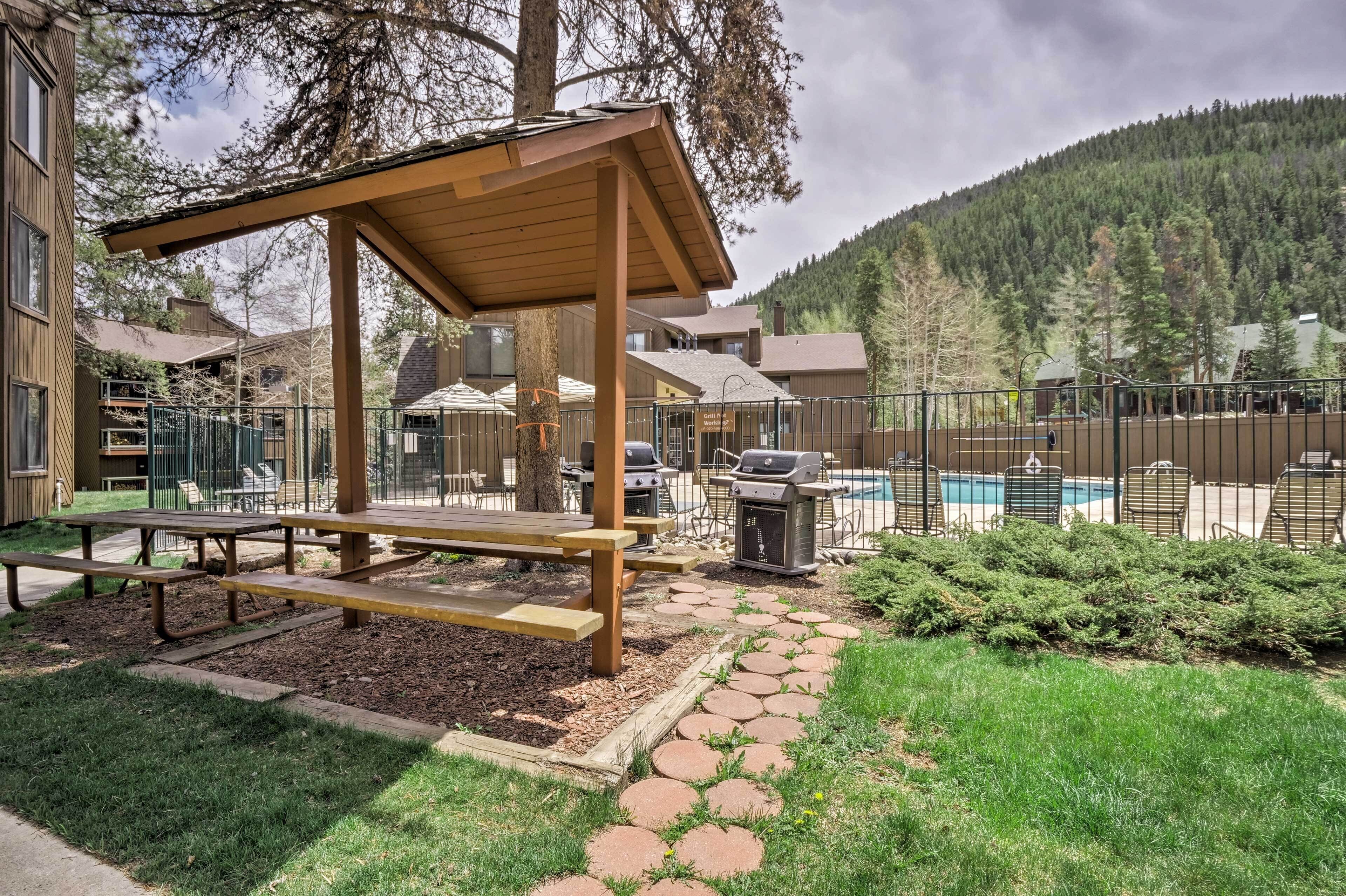 A picnic area with a wooden bench and picnic table, surrounded by a fenced pool area with a barbecue grill.