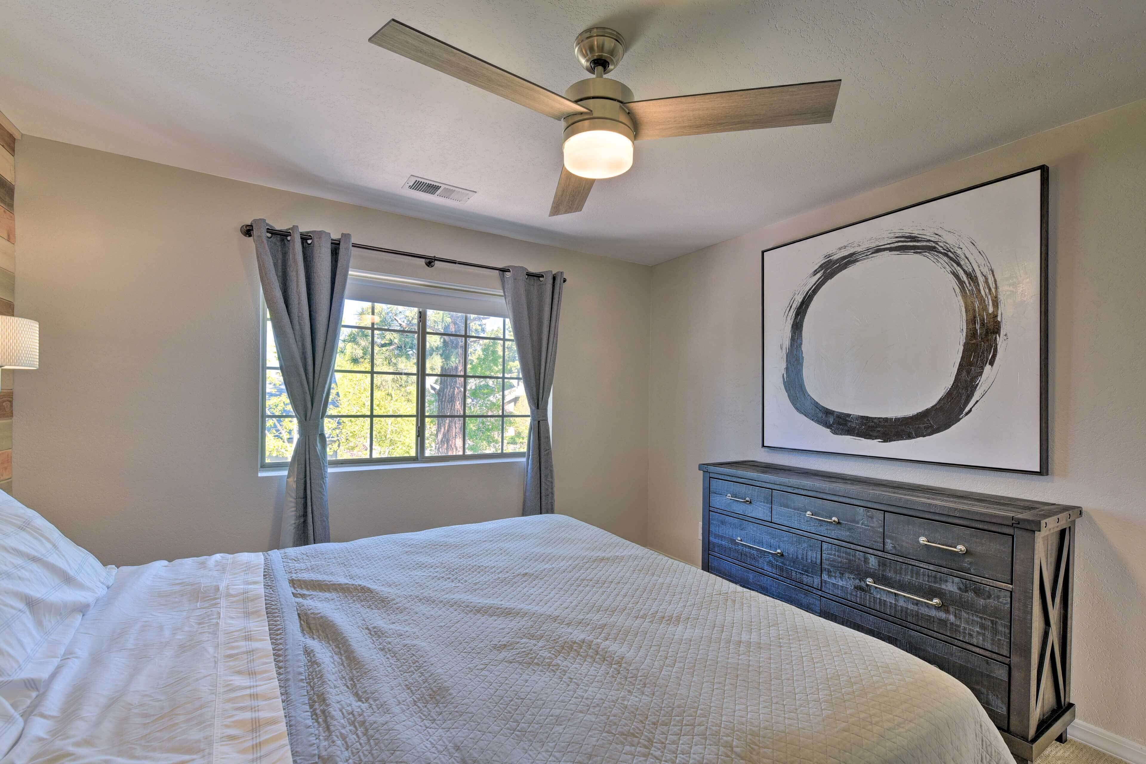 A bedroom with a bed, a ceiling fan, a window with curtains, a dresser, and a painting on the wall.