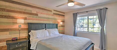 A bedroom with a wooden slat wall, a bed with white bedding, a nightstand with a clock, a ceiling fan, and a window with curtains.