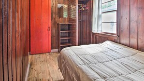 Ein Schlafzimmer mit Holzwänden, einem roten Kleiderschrank, einem Bett mit beige farbenem Überwurf und einem Fenster mit Blick.