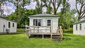 Eine kleine, eingeschossige Hütte mit Veranda und einer Holzterrasse, umgeben von üppigem Grün und Bäumen.